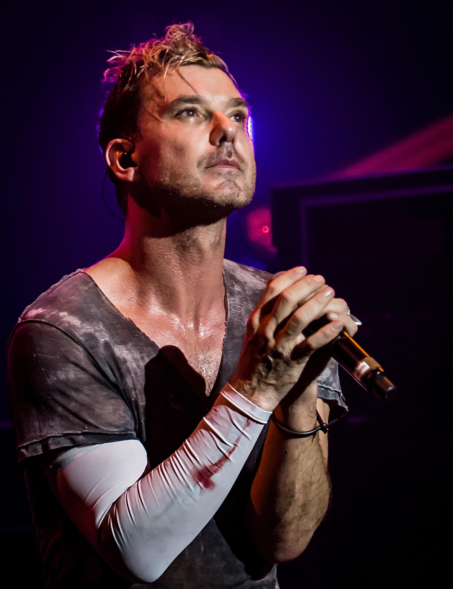 A singer wearing a grey shirt, with a white sleeve bearing a blood stain, clutches a microphone against a dark blue background.