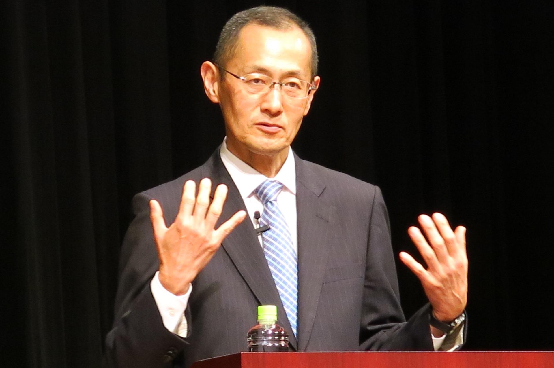 A man in a suit stands at a podium against a black background.