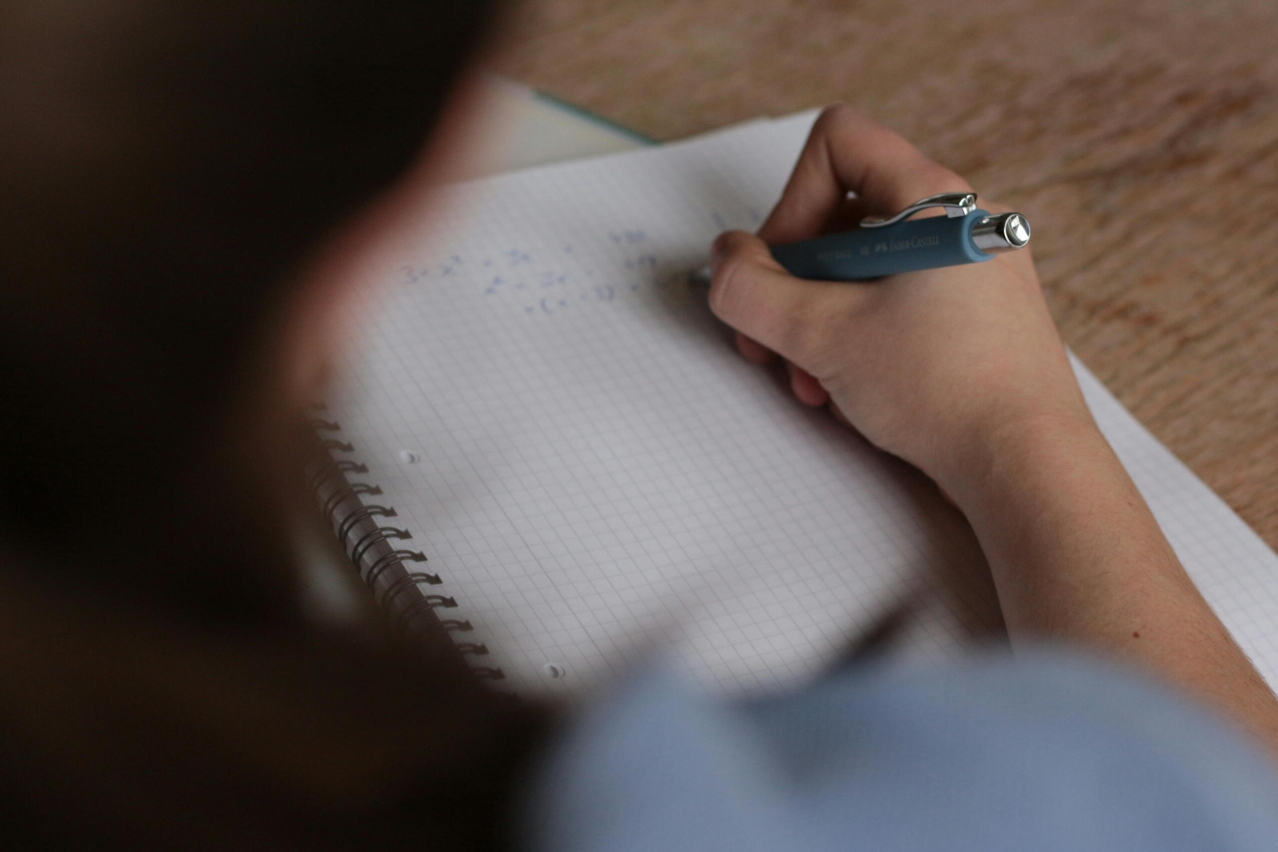 A girl sits at a desk writing in a math notebook, focusing intently as she works through equations with a pencil.